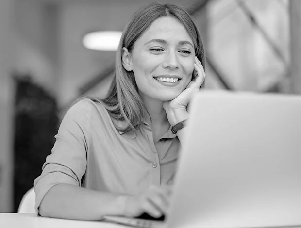 Grayscale close-up of a middle-aged woman with shoulder-length light brown hair, seated at a table and looking at a laptop. She wears a light-colored long-sleeve shirt, rests her right hand on her cheek, and smiles gently. The background is a softly blurred, light-toned room suggesting a casual workspace. - Blog in Atlanta, GA