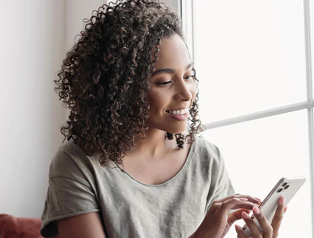 A medium-brown skinned woman with curly dark brown hair sits near a bright window, looking down at a light-colored smartphone with a smile. She wears a short-sleeved, light gray/taupe t-shirt. A reddish-brown couch or chair is partially visible in the lower left, and the background is neutral. - Reviews in Atlanta, GA